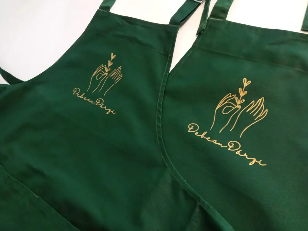 Two green aprons with a gold Debesu Dārzi logo