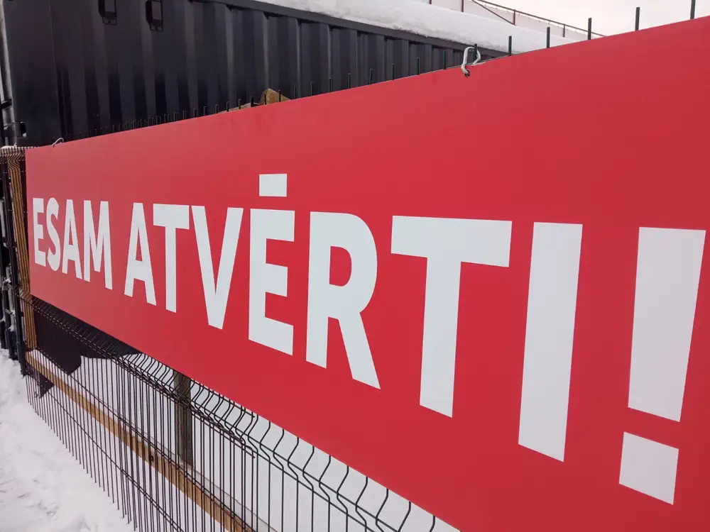 Red banner with the white text "Esam atvērti" on a fence in winter