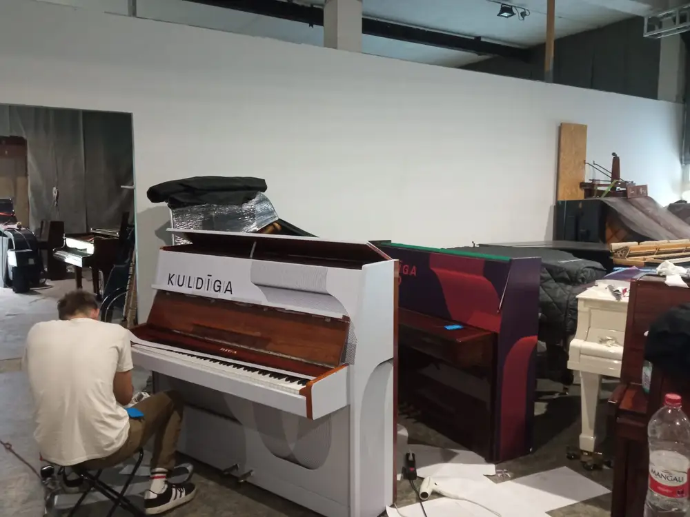 A worker wrapping an upright piano in white film with the word Kuldīga