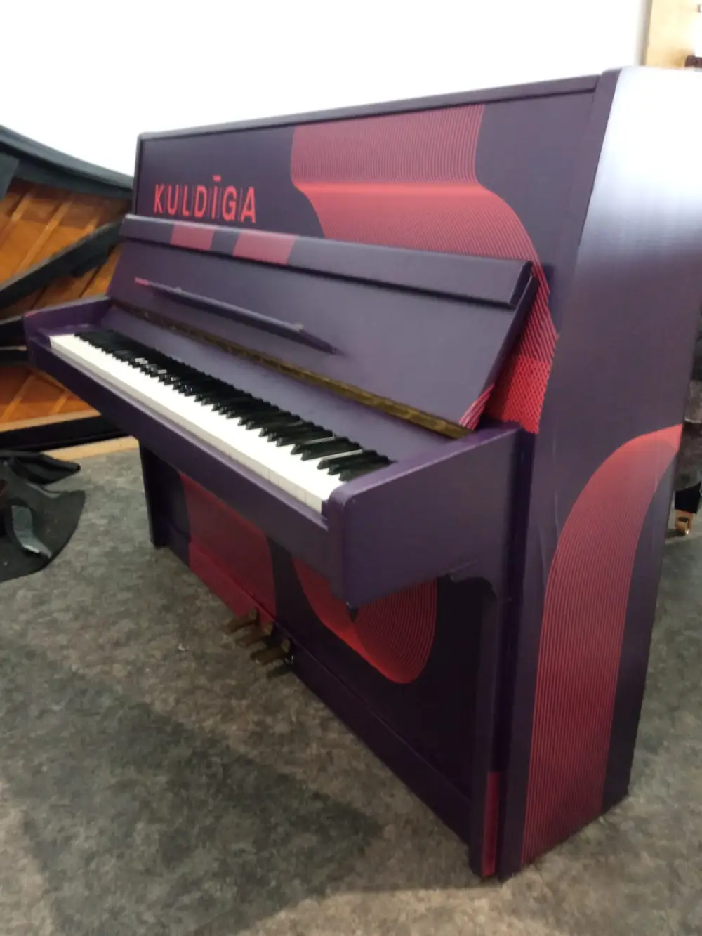 Upright piano wrapped in a purple and red pattern with the word Kuldīga