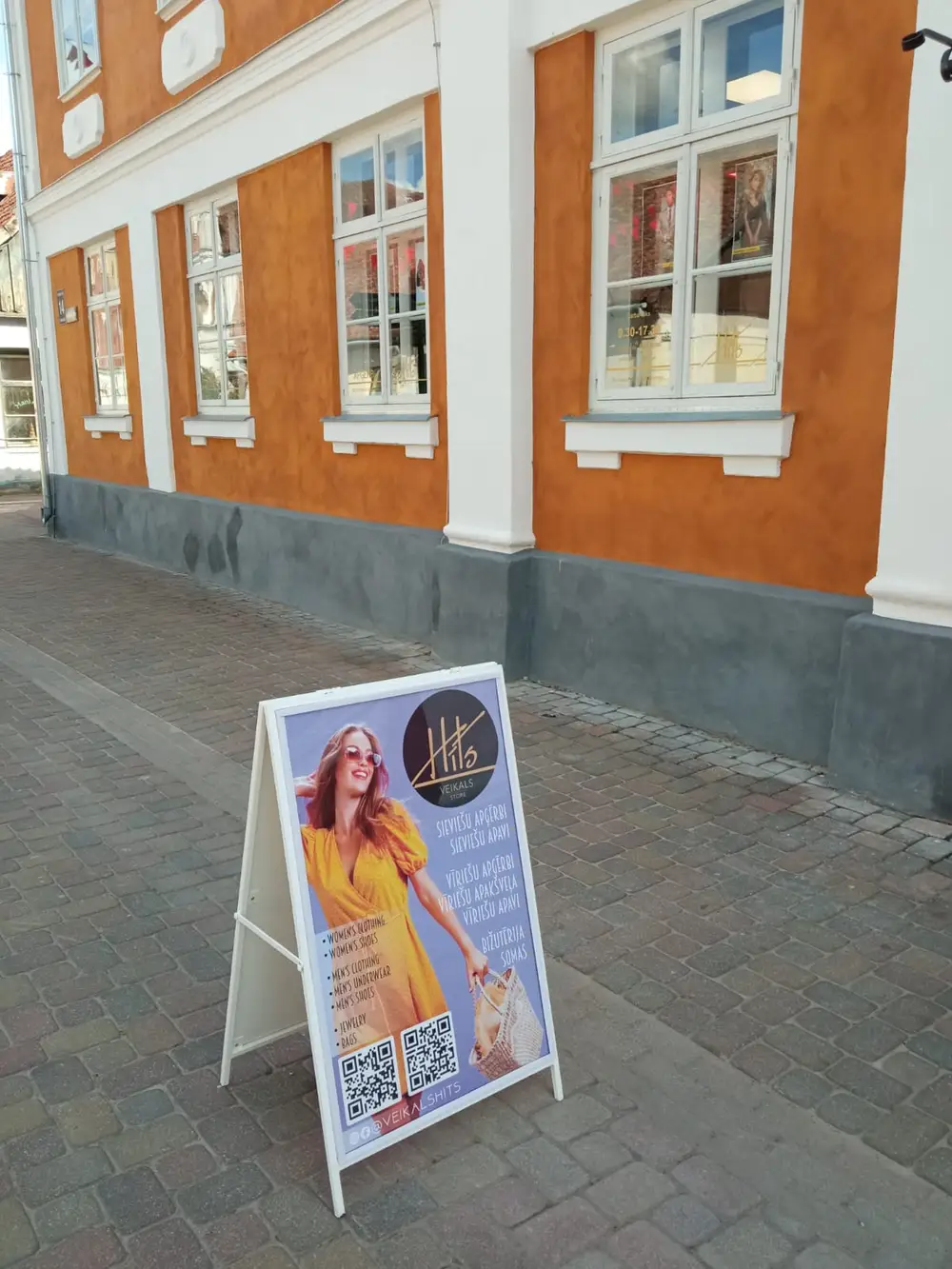 White pavement sign with a Hits shop poster on the paving by a building