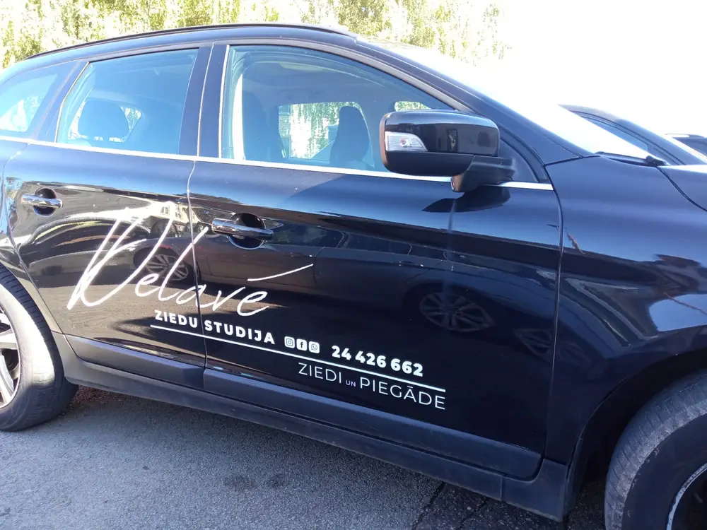 Black car side doors with white Delavé ziedu studija lettering and a phone number