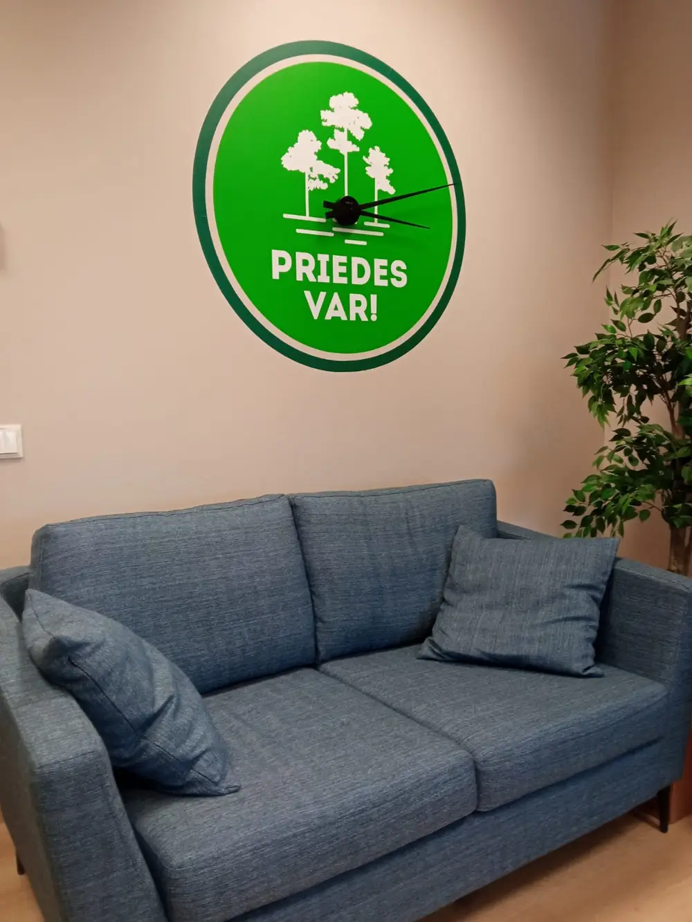 Green round wall clock with trees and the text "Priedes var!" above a sofa
