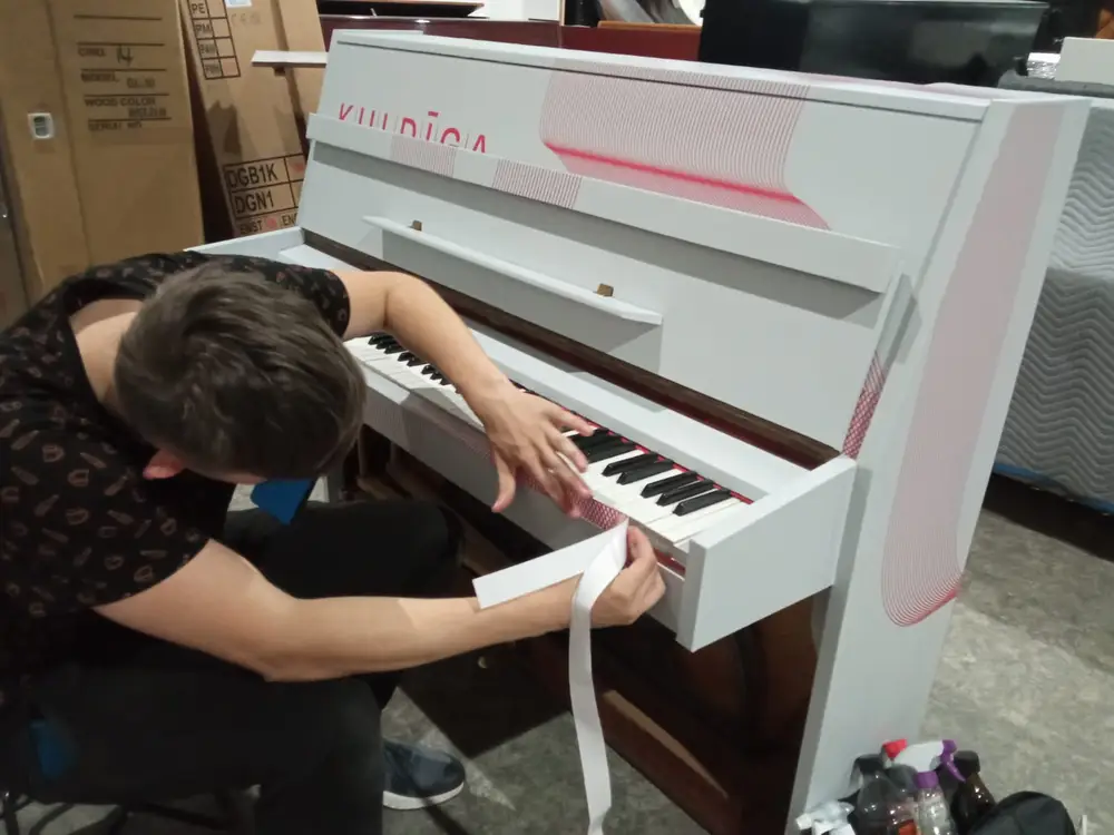 A worker applying film along the keyboard edge of a white upright piano