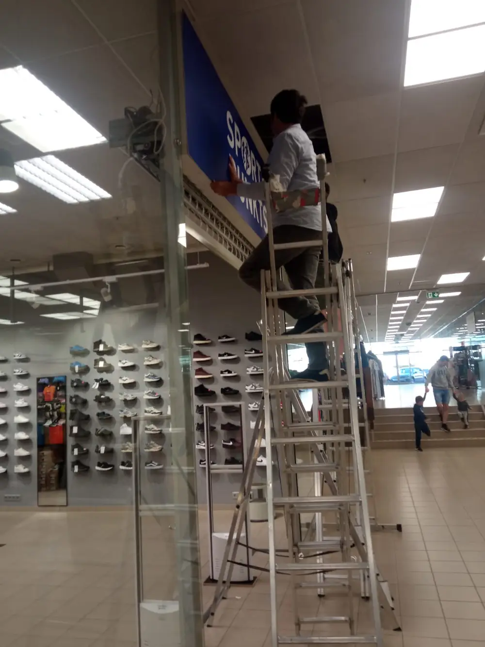 Installer on a ladder fitting the blue Sporta Punkts sign in a shopping centre