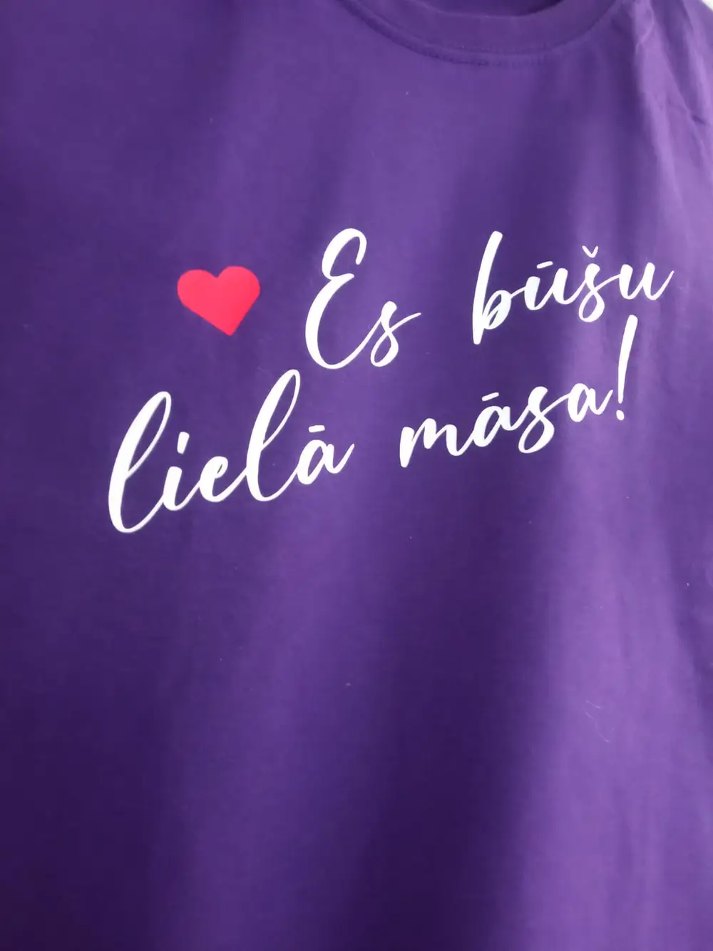 Purple T-shirt with the white lettering "Es būšu lielā māsa" and a red heart