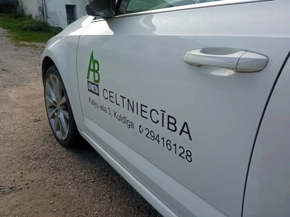 White car door with a green AB logo, the black word "Celtniecība", an address in Kuldīga and a phone number