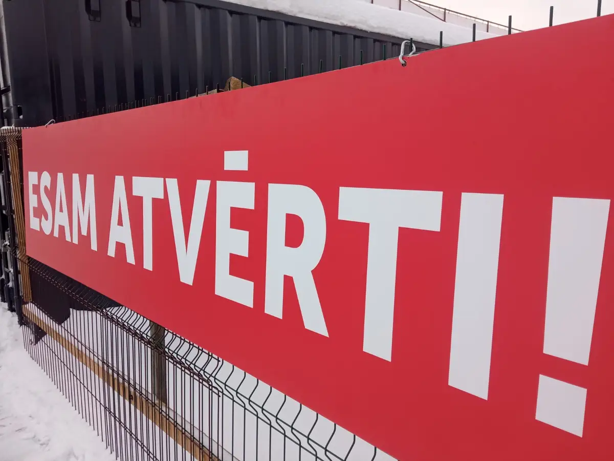 Red PVC banner with the white words "Esam atvērti!" on a metal fence in winter
