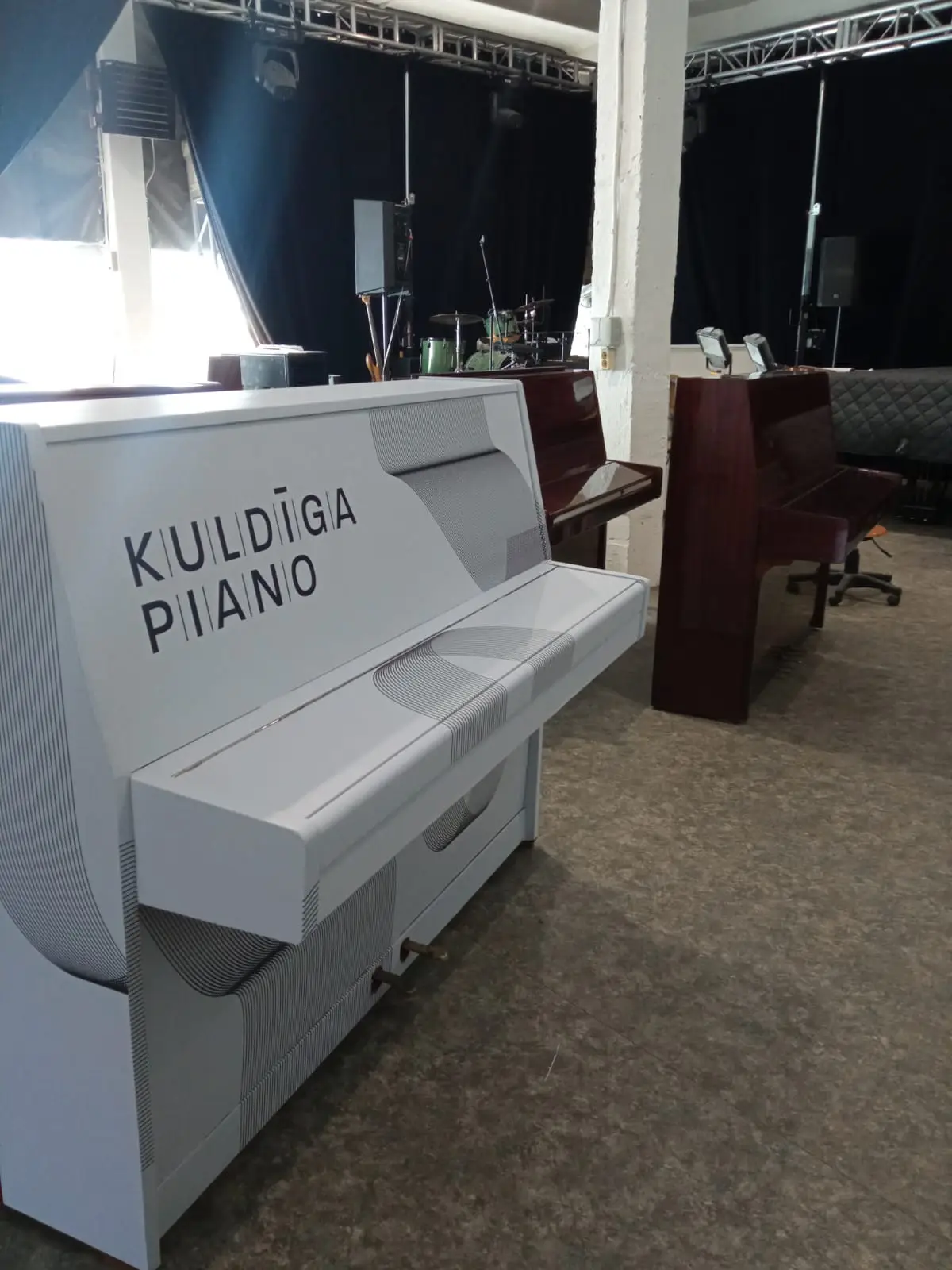 White piano with black "Kuldīga Piano" lettering and a fine line pattern on the body