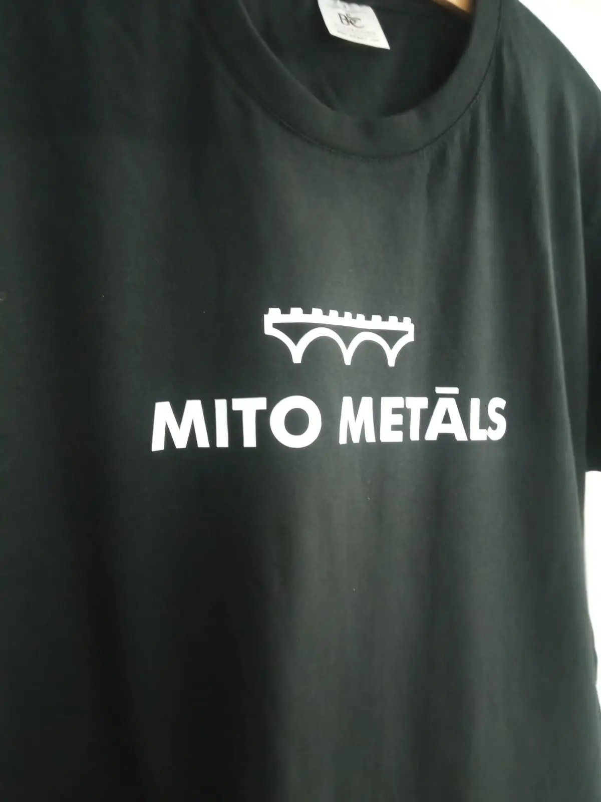 Dark T-shirt with a white "Mito Metāls" logo and a bridge silhouette