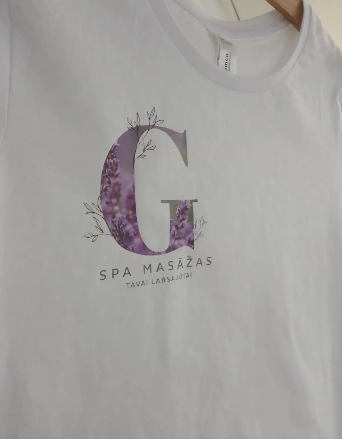 White T-shirt with a letter G filled with a lavender pattern and the text "Spa masāžas, tavai labsajūtai"